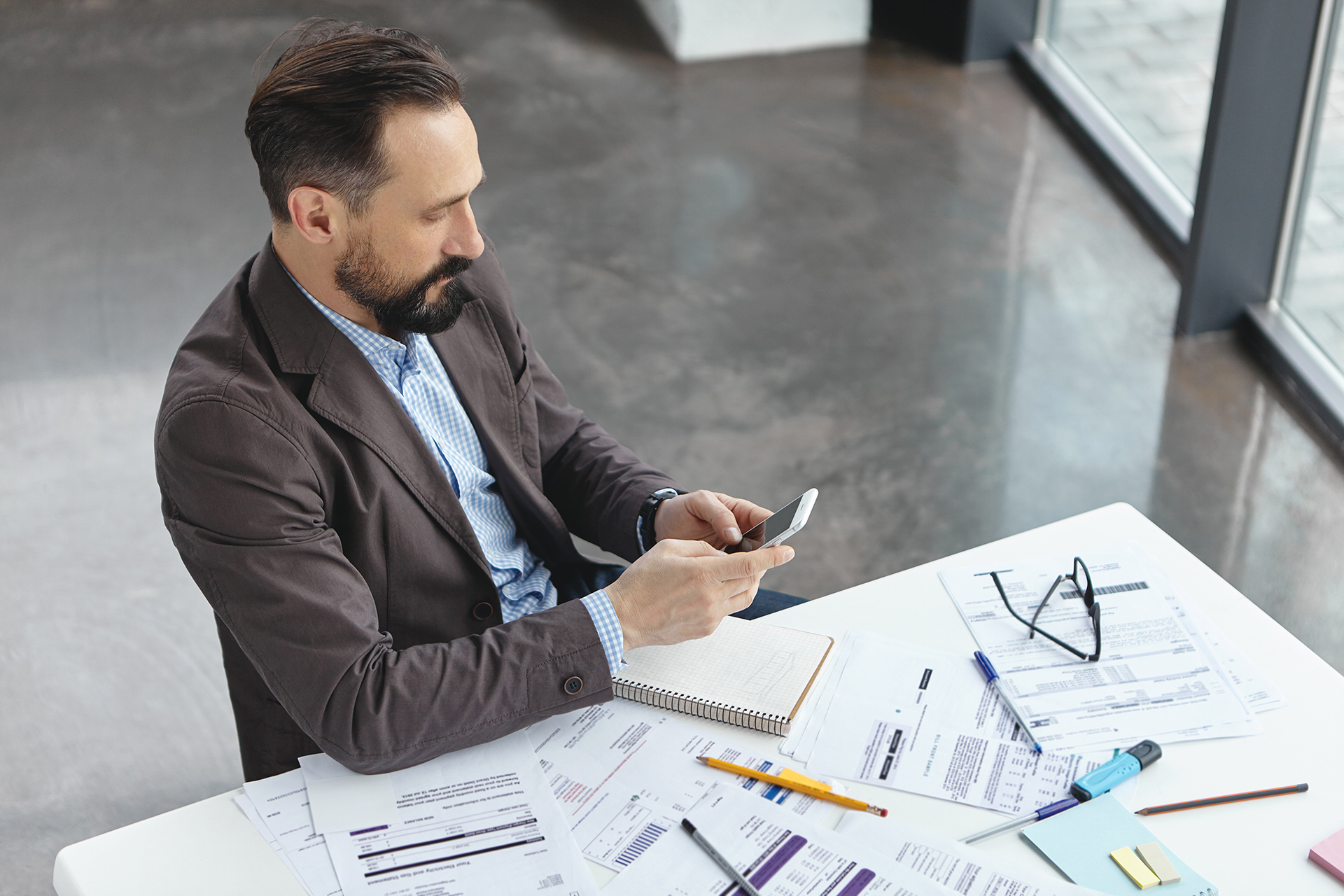 Top view of concentrated middle aged businessman dressed in formal suit, sits at work place, surrounded with many documents, holds smart phone, reads news online or makes transactions, check email